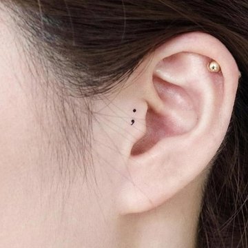 Minimalistisches Tattoo eines Semikolons hinter dem Ohr